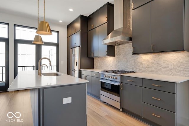 kitchen with decorative light fixtures, light hardwood / wood-style flooring, appliances with stainless steel finishes, an island with sink, and wall chimney range hood