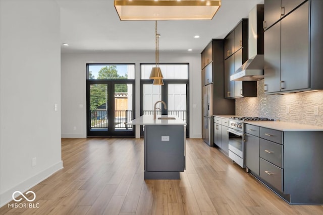 kitchen featuring wall chimney range hood, an island with sink, stainless steel appliances, and light hardwood / wood-style floors