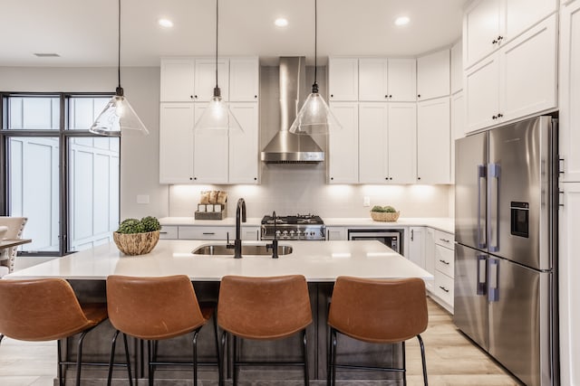 kitchen featuring high quality fridge, light wood-type flooring, gas range oven, wall chimney range hood, and a kitchen island with sink