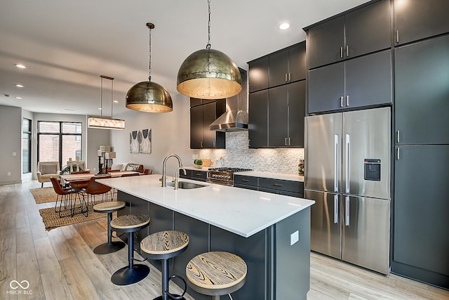 kitchen featuring decorative light fixtures, stainless steel appliances, light countertops, a kitchen island with sink, and wall chimney range hood