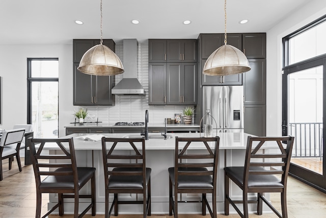 kitchen with a center island with sink, wall chimney range hood, stainless steel fridge with ice dispenser, and light wood-type flooring