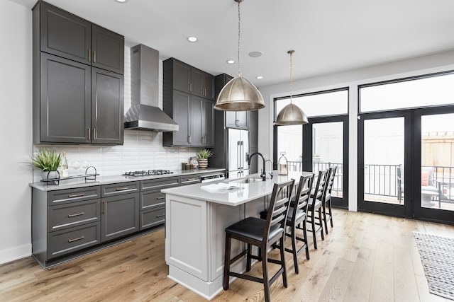 kitchen featuring light hardwood / wood-style floors, an island with sink, backsplash, wall chimney exhaust hood, and appliances with stainless steel finishes