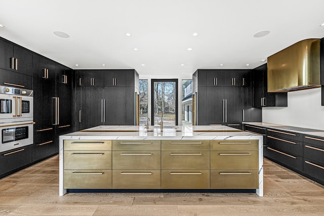 kitchen featuring stainless steel double oven, wall chimney range hood, light wood-type flooring, and light stone countertops