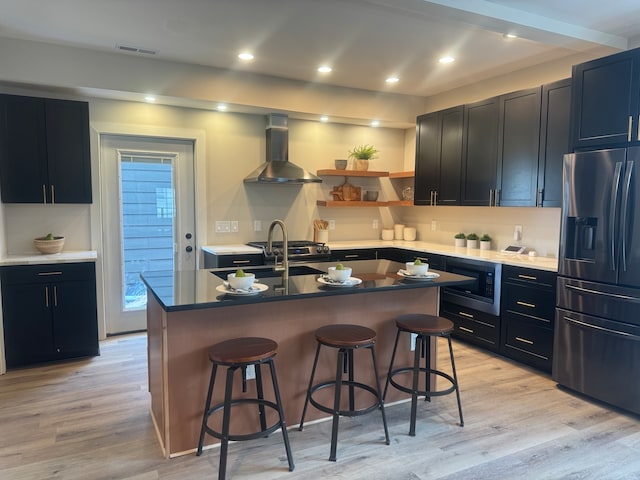 kitchen featuring black microwave, wall chimney range hood, light hardwood / wood-style floors, stainless steel fridge, and a kitchen island with sink