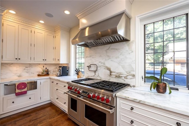 kitchen with dark wood-type flooring, range with two ovens, light stone countertops, wall chimney exhaust hood, and backsplash