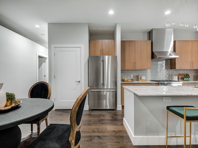 kitchen featuring backsplash, dark wood-type flooring, wall chimney range hood, stainless steel refrigerator, and pendant lighting