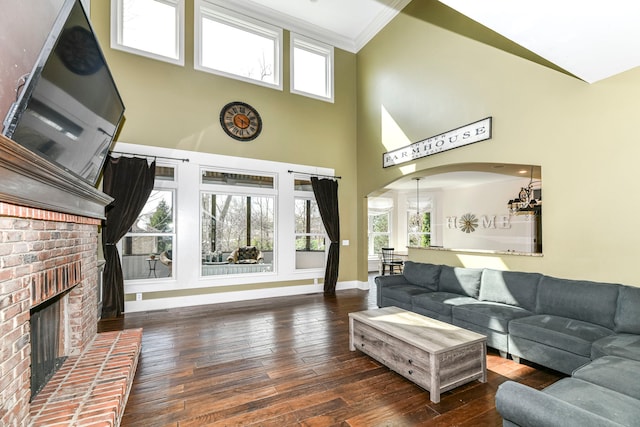 living room featuring a high ceiling, dark wood-type flooring, a notable chandelier, a brick fireplace, and crown molding