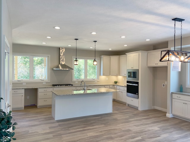 kitchen featuring a center island with sink, hanging light fixtures, wall chimney exhaust hood, appliances with stainless steel finishes, and white cabinetry