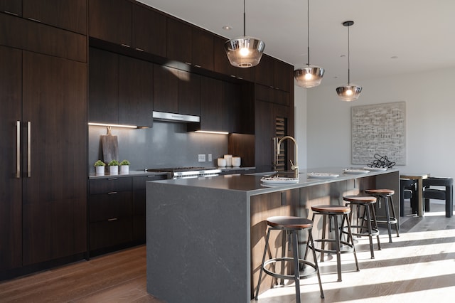 kitchen with light wood-type flooring, backsplash, sink, a center island with sink, and hanging light fixtures