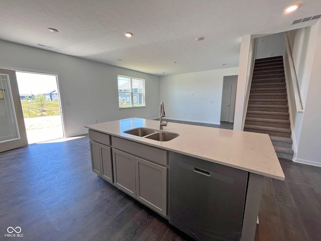 kitchen with stainless steel dishwasher, sink, dark wood-type flooring, an island with sink, and light stone counters