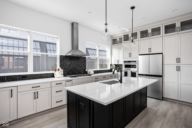 kitchen featuring appliances with stainless steel finishes, sink, wall chimney exhaust hood, an island with sink, and pendant lighting