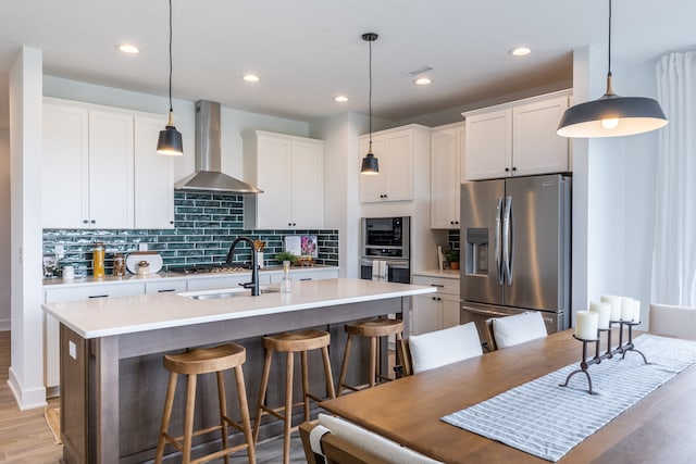 kitchen with wall chimney exhaust hood, stainless steel appliances, an island with sink, and decorative light fixtures