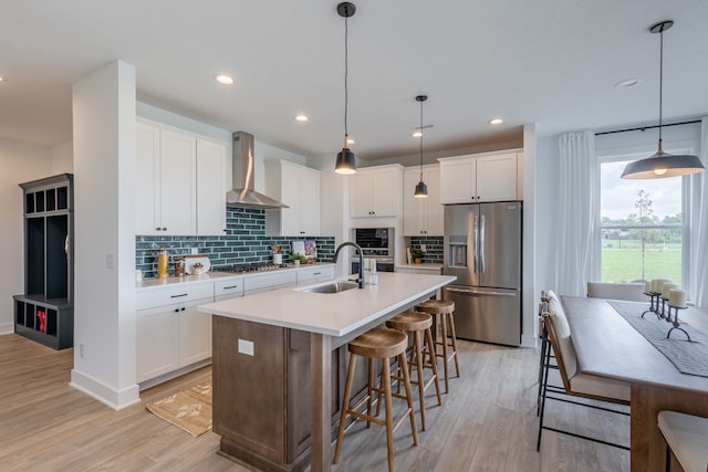 kitchen with wall chimney exhaust hood, appliances with stainless steel finishes, decorative light fixtures, and a kitchen island with sink