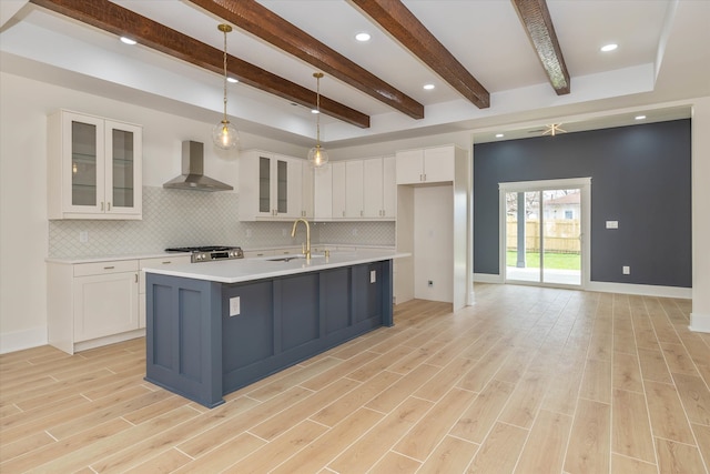 kitchen with sink, white cabinetry, an island with sink, wall chimney range hood, and pendant lighting