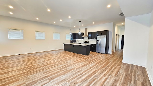 kitchen with appliances with stainless steel finishes, a kitchen island with sink, wall chimney range hood, light hardwood / wood-style floors, and hanging light fixtures