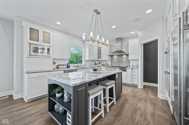 kitchen featuring a center island, wall chimney range hood, hanging light fixtures, light stone countertops, and white cabinetry