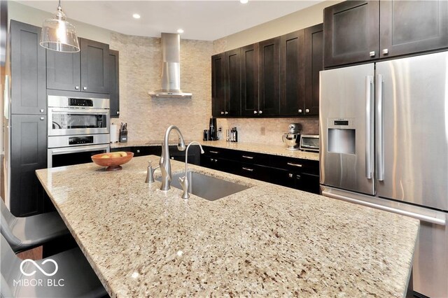 kitchen with stainless steel appliances, light stone countertops, wall chimney exhaust hood, sink, and pendant lighting