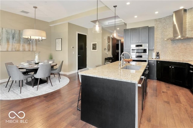 kitchen featuring wall chimney range hood, backsplash, a center island with sink, hardwood / wood-style floors, and sink