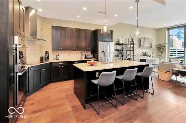 kitchen featuring an island with sink, light wood-type flooring, backsplash, wall chimney exhaust hood, and appliances with stainless steel finishes