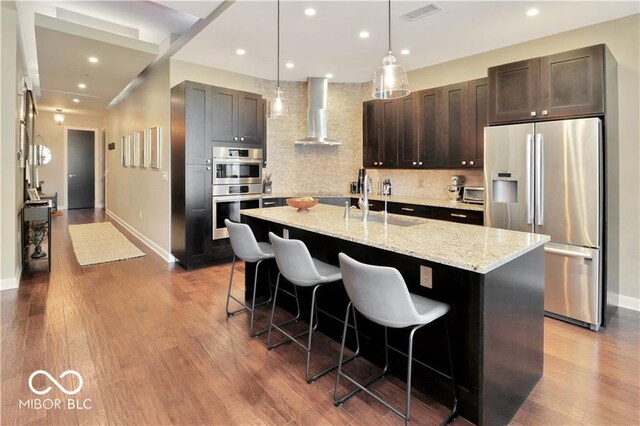 kitchen featuring wall chimney range hood, backsplash, a center island with sink, wood-type flooring, and appliances with stainless steel finishes