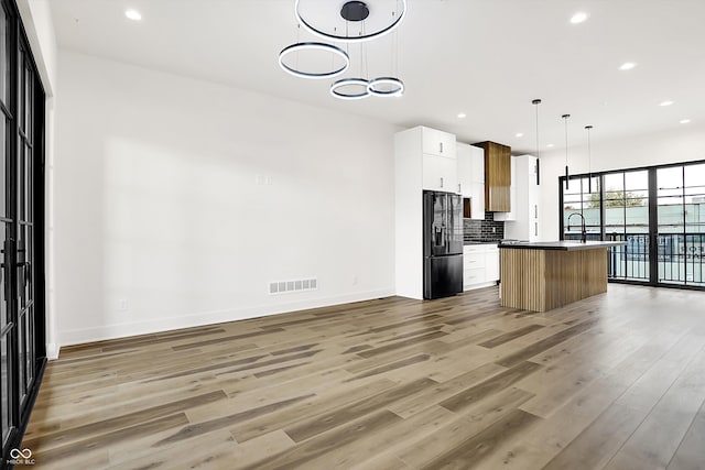 kitchen with white cabinetry, light hardwood / wood-style flooring, an island with sink, decorative light fixtures, and black fridge with ice dispenser