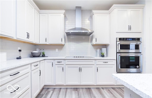 kitchen featuring white cabinetry, light hardwood / wood-style floors, tasteful backsplash, double oven, and wall chimney range hood