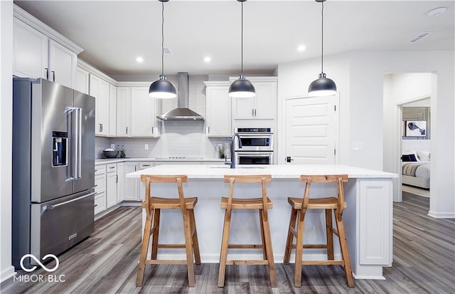 kitchen with white cabinets, decorative backsplash, wall chimney range hood, a kitchen island with sink, and appliances with stainless steel finishes