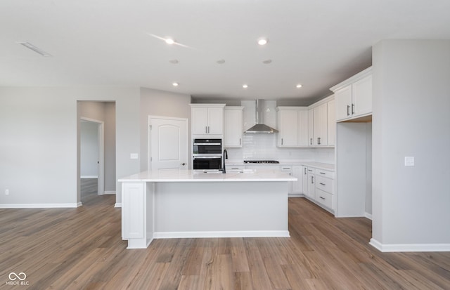 kitchen with light hardwood / wood-style flooring, an island with sink, wall chimney range hood, decorative backsplash, and white cabinetry