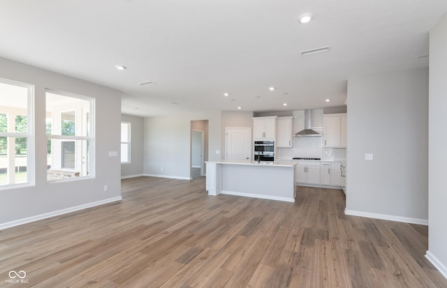 kitchen with wall chimney exhaust hood, an island with sink, decorative backsplash, light wood-type flooring, and white cabinets