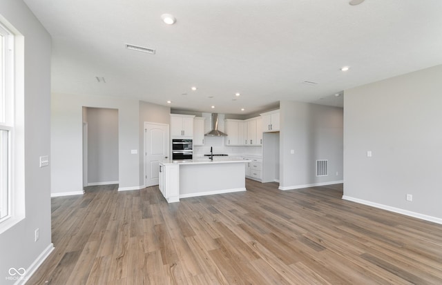 kitchen with a center island with sink, light hardwood / wood-style floors, white cabinets, wall chimney range hood, and sink