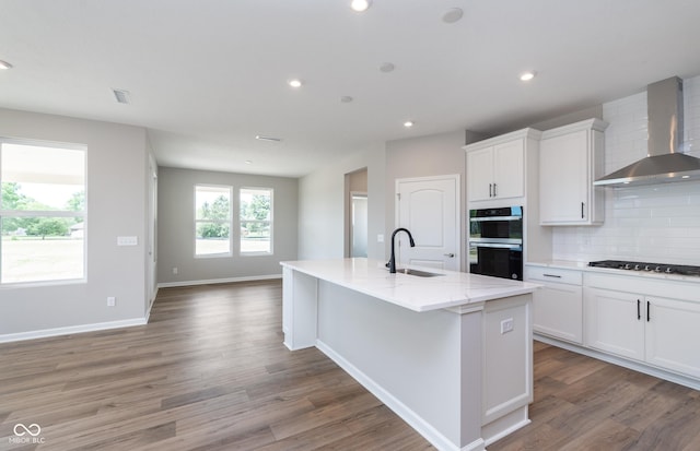 kitchen featuring sink, wall chimney exhaust hood, an island with sink, light stone countertops, and black appliances