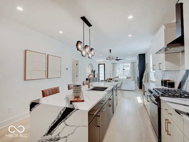 kitchen with a sink, wall chimney range hood, light wood-type flooring, black range with gas stovetop, and an island with sink