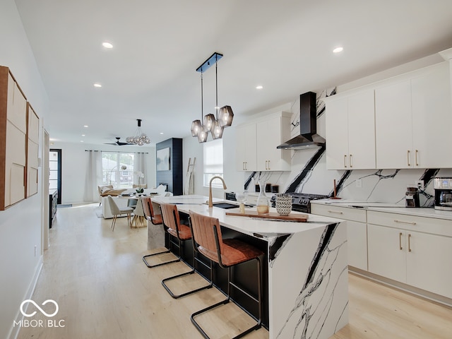 kitchen featuring light wood finished floors, backsplash, white cabinetry, wall chimney range hood, and an island with sink