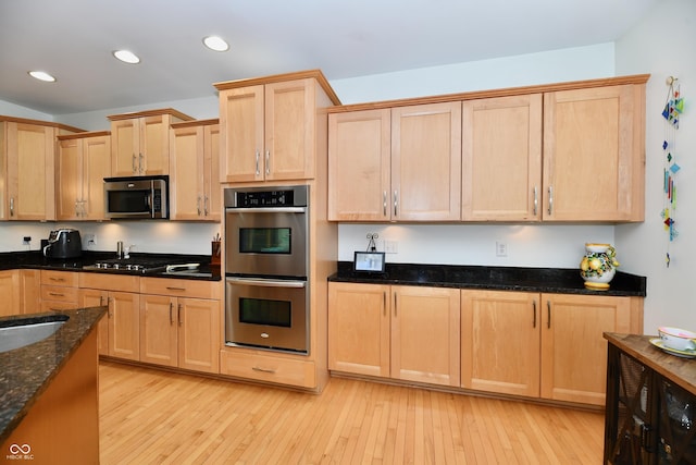 kitchen with light wood-type flooring, appliances with stainless steel finishes, dark stone counters, and light brown cabinets