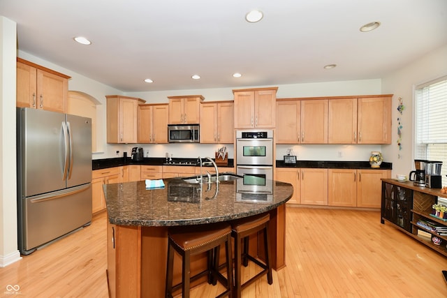 kitchen featuring appliances with stainless steel finishes, light hardwood / wood-style floors, dark stone counters, and a kitchen island with sink