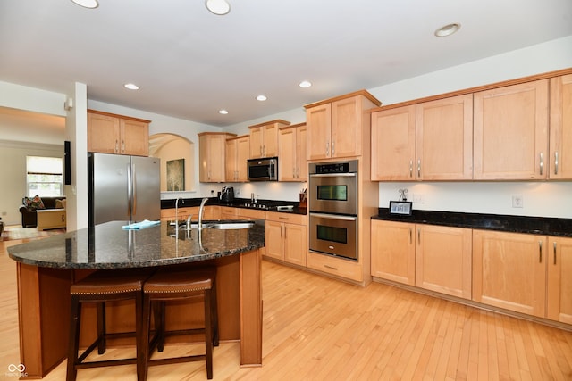 kitchen featuring light hardwood / wood-style floors, an island with sink, appliances with stainless steel finishes, dark stone countertops, and sink