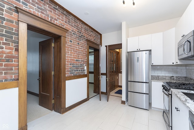 kitchen featuring light stone countertops, appliances with stainless steel finishes, decorative backsplash, brick wall, and white cabinetry