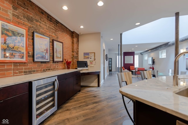 bar featuring beverage cooler, light wood-type flooring, light stone counters, dark brown cabinetry, and brick wall