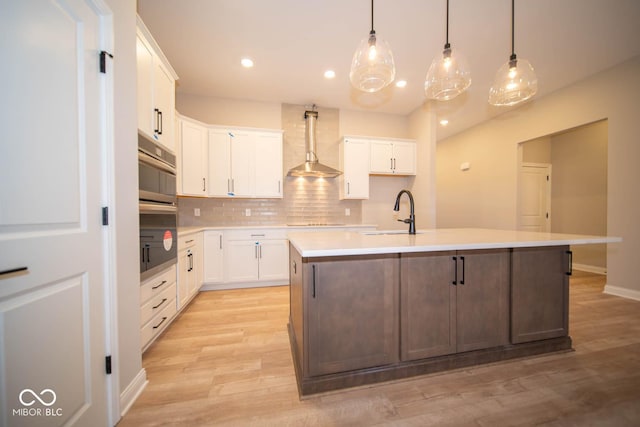 kitchen with sink, white cabinetry, an island with sink, and wall chimney range hood