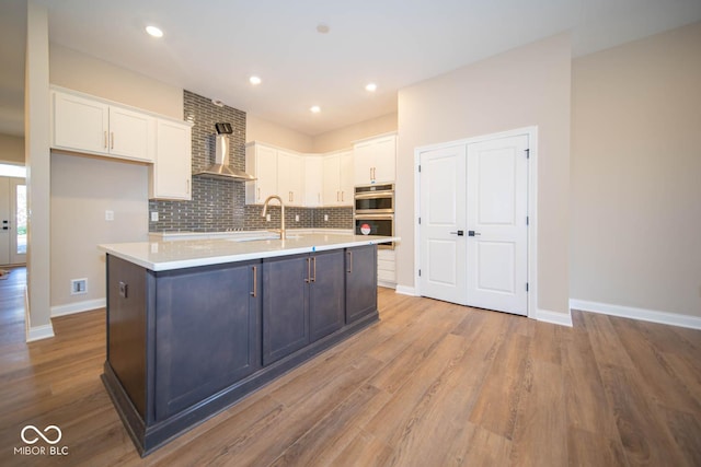 kitchen with white cabinetry, wall chimney exhaust hood, stainless steel double oven, light hardwood / wood-style floors, and a kitchen island with sink