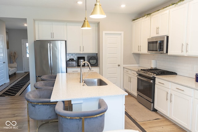 kitchen featuring appliances with stainless steel finishes, an island with sink, light wood-type flooring, decorative light fixtures, and sink