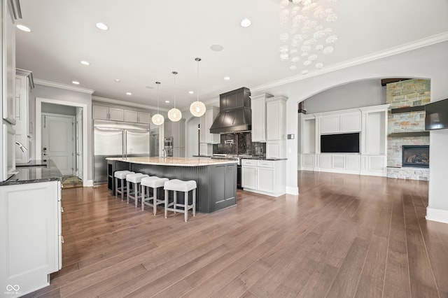 kitchen with hardwood / wood-style floors, a stone fireplace, custom exhaust hood, a large island, and built in refrigerator