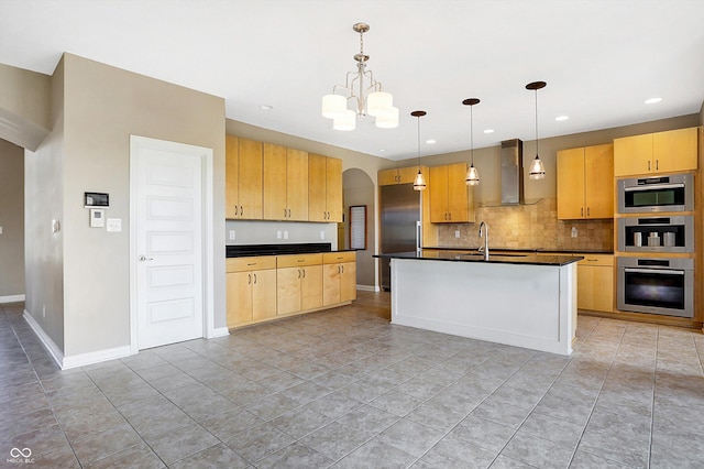 kitchen featuring a kitchen island with sink, light brown cabinetry, wall chimney exhaust hood, decorative light fixtures, and backsplash