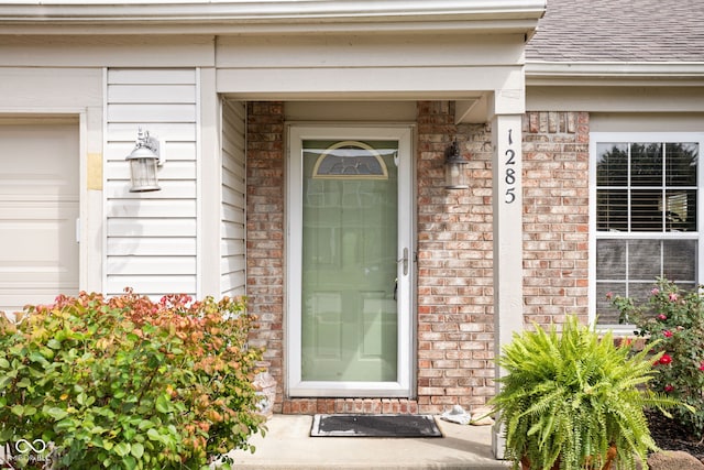 doorway to property featuring brick siding, an attached garage, and roof with shingles