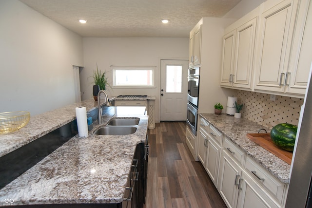 kitchen featuring double oven, sink, light stone counters, dark wood-type flooring, and a center island with sink