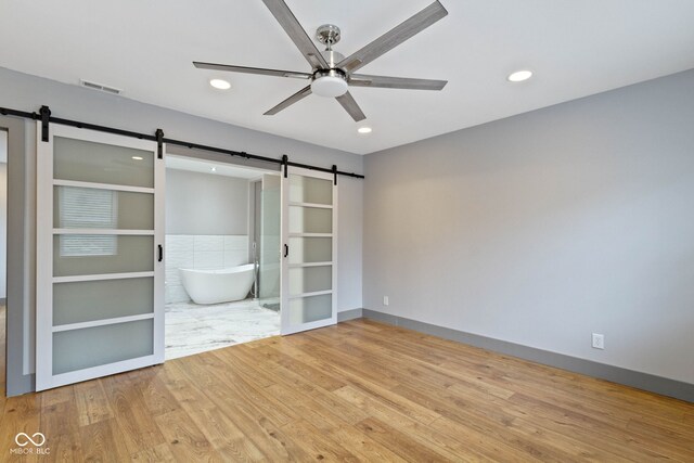 unfurnished bedroom with light hardwood / wood-style floors, a barn door, ceiling fan, and ensuite bathroom