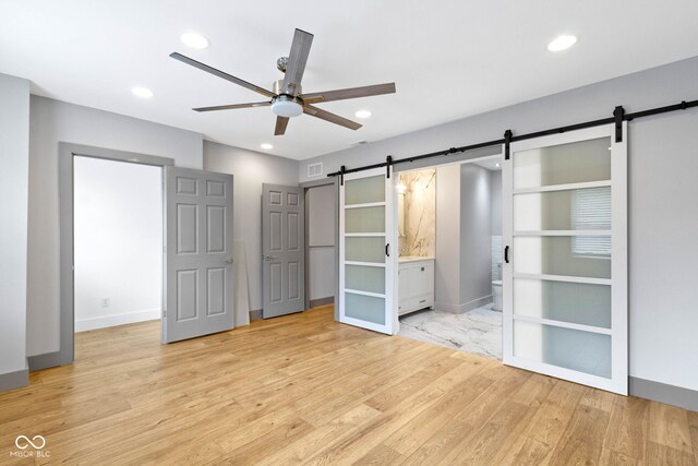 unfurnished bedroom with ceiling fan, ensuite bathroom, light hardwood / wood-style floors, and a barn door