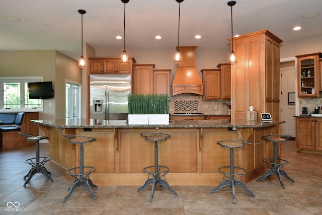 kitchen with brown cabinets, stainless steel refrigerator with ice dispenser, a breakfast bar area, tasteful backsplash, and custom range hood