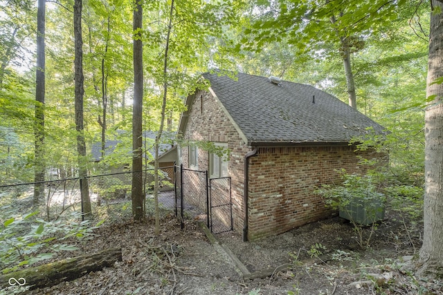 view of side of home with brick siding, a shingled roof, a gate, fence, and a wooded view