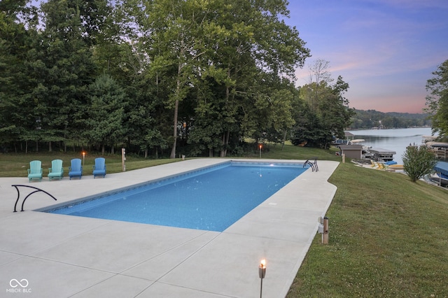 pool at dusk with a water view, a patio area, and a lawn